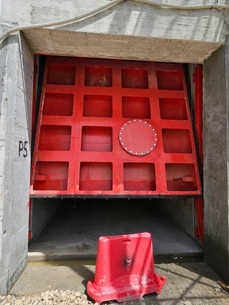Porte sectionnelle rouge à raidisseurs et trappe boulonnée, partiellement relevée, en galerie de tunnel
